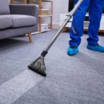 A professional carpet cleaning technician in blue overalls and protective shoe covers uses a steam cleaning wand on a grey ribbed carpet in a living room.