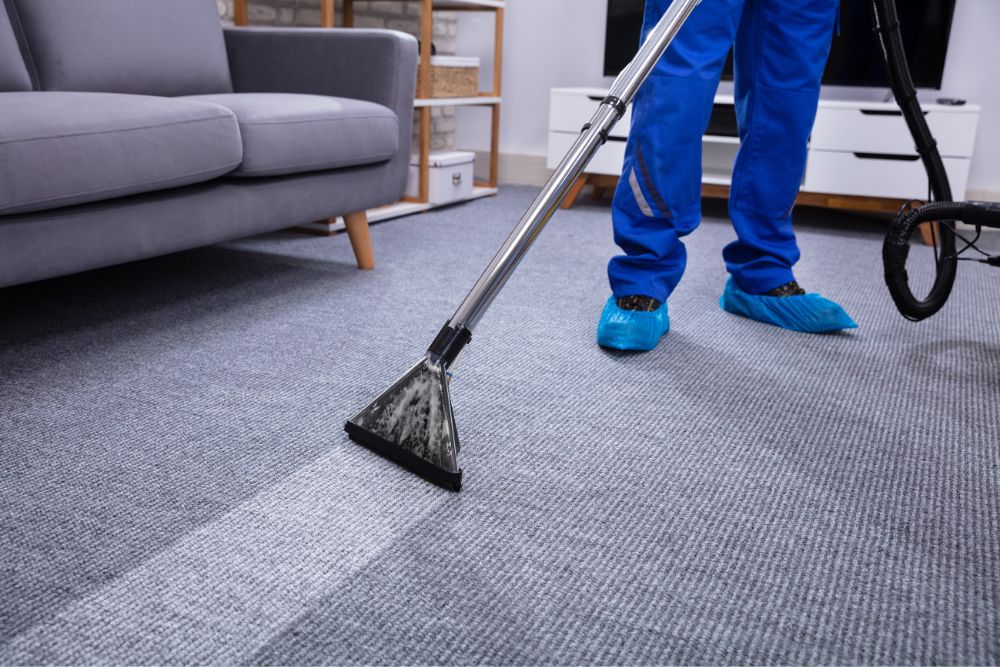 A professional carpet cleaning technician in blue overalls and protective shoe covers uses a steam cleaning wand on a grey ribbed carpet in a living room.