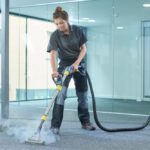 A professional female cleaner in a grey polo shirt and dark work trousers uses a steam cleaning wand on a grey commercial carpet.