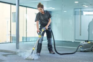 A professional female cleaner in a grey polo shirt and dark work trousers uses a steam cleaning wand on a grey commercial carpet.