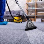 A professional carpet cleaner in a blue uniform and blue shoe covers uses a steam cleaning wand on a grey textured carpet in a modern living room.