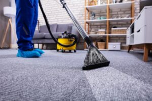 A professional carpet cleaner in a blue uniform and blue shoe covers uses a steam cleaning wand on a grey textured carpet in a modern living room.